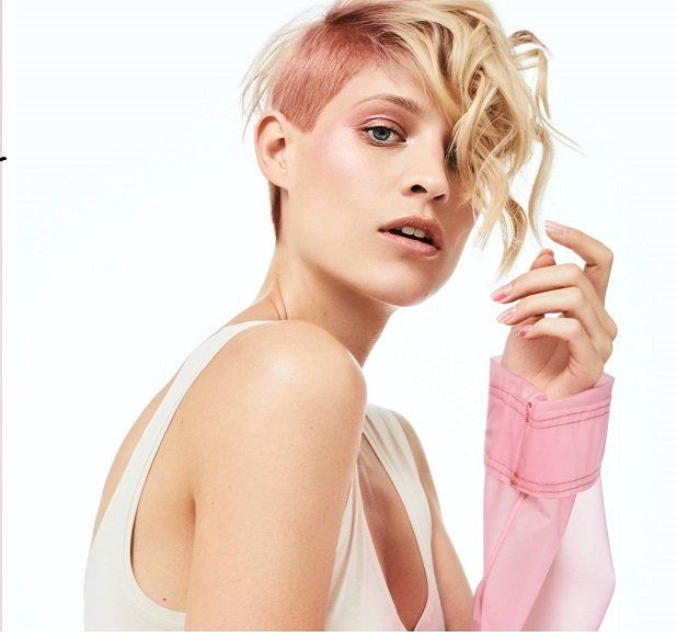 Una mujer con cabello rubio corto lleva una camisa rosa.