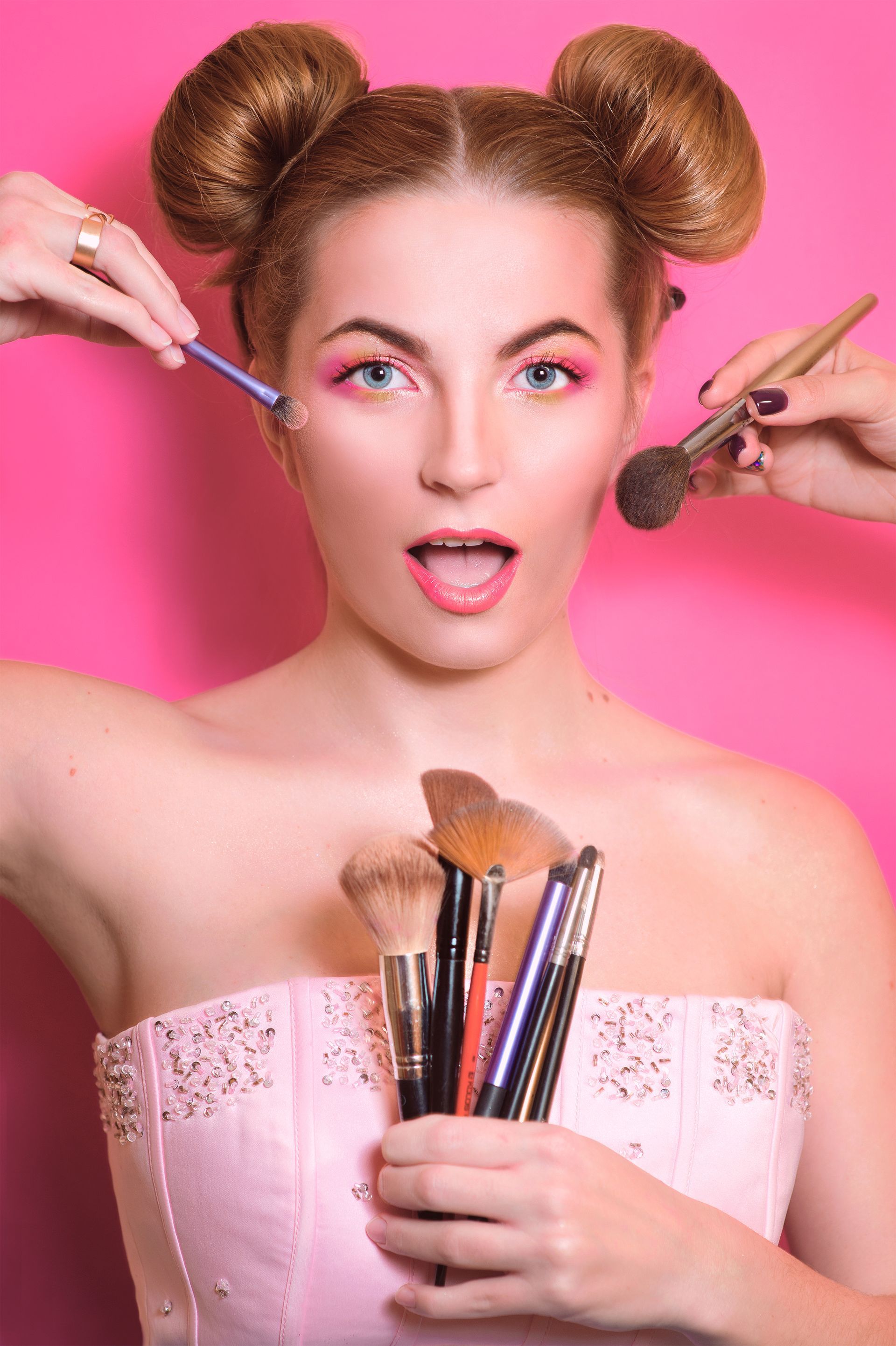 Mujer recibiendo maquillaje aplicado con múltiples manos, piel clara, fondo rosa.