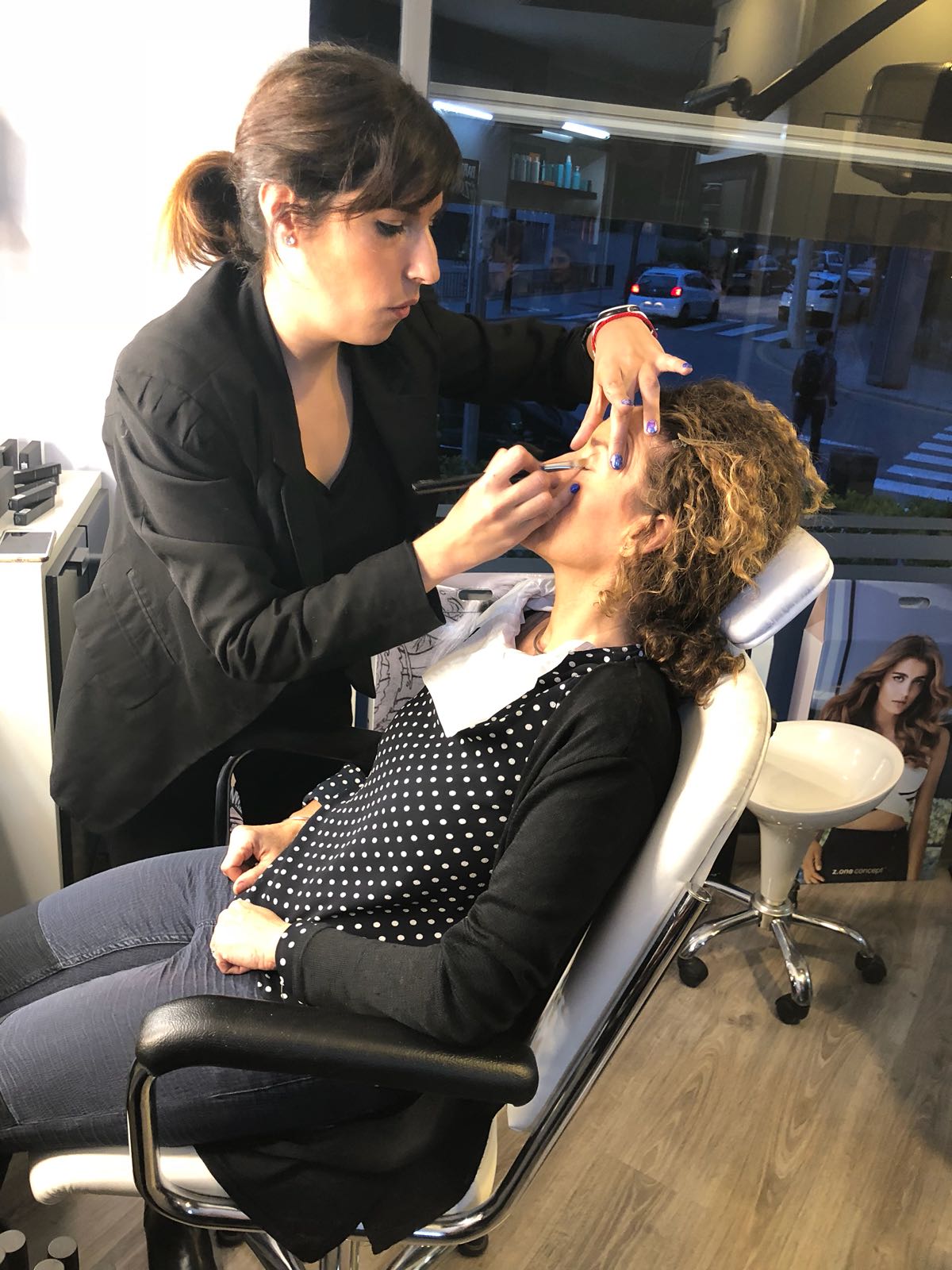 Una mujer está sentada en una silla y otra mujer la maquilla.