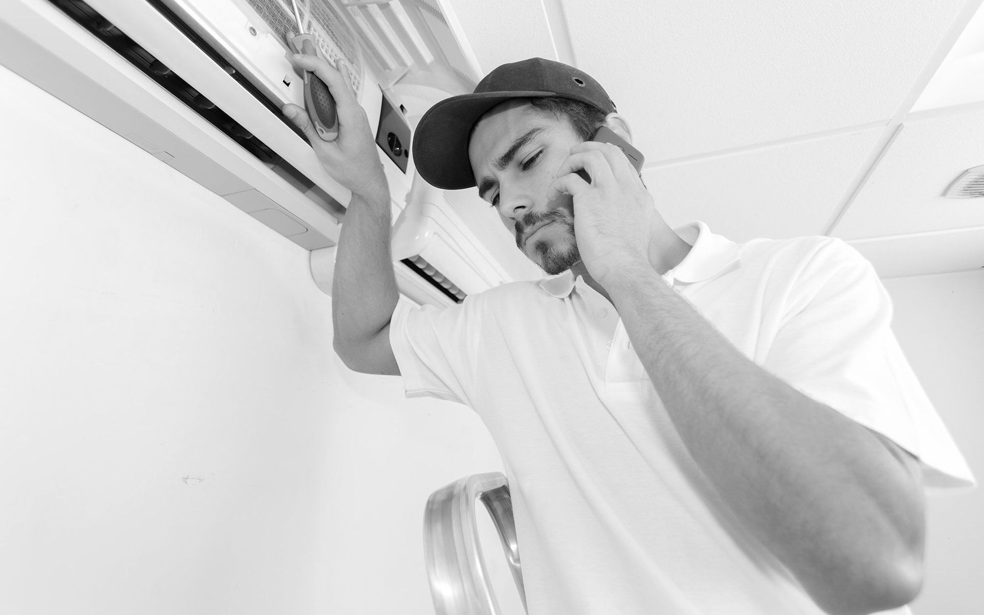 Technician inspecting an air conditioner indoors, holding a phone while adjusting the unit.