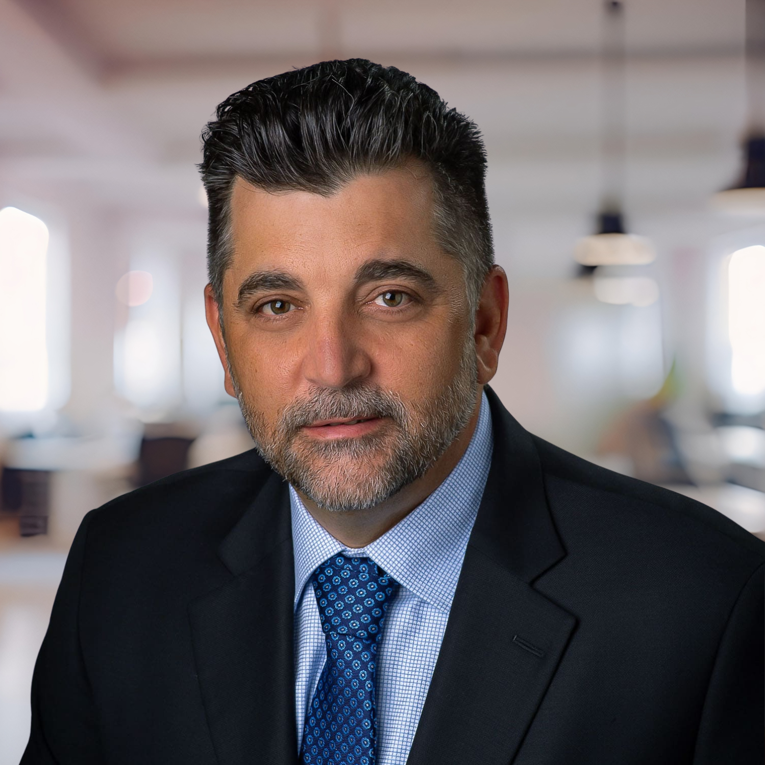 Portrait of a man in a dark suit and blue tie, posing against a softly blurred office background