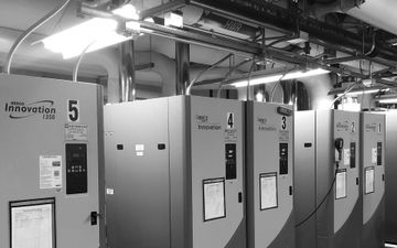 Row of large industrial electrical cabinets in a utility room with overhead piping and cables