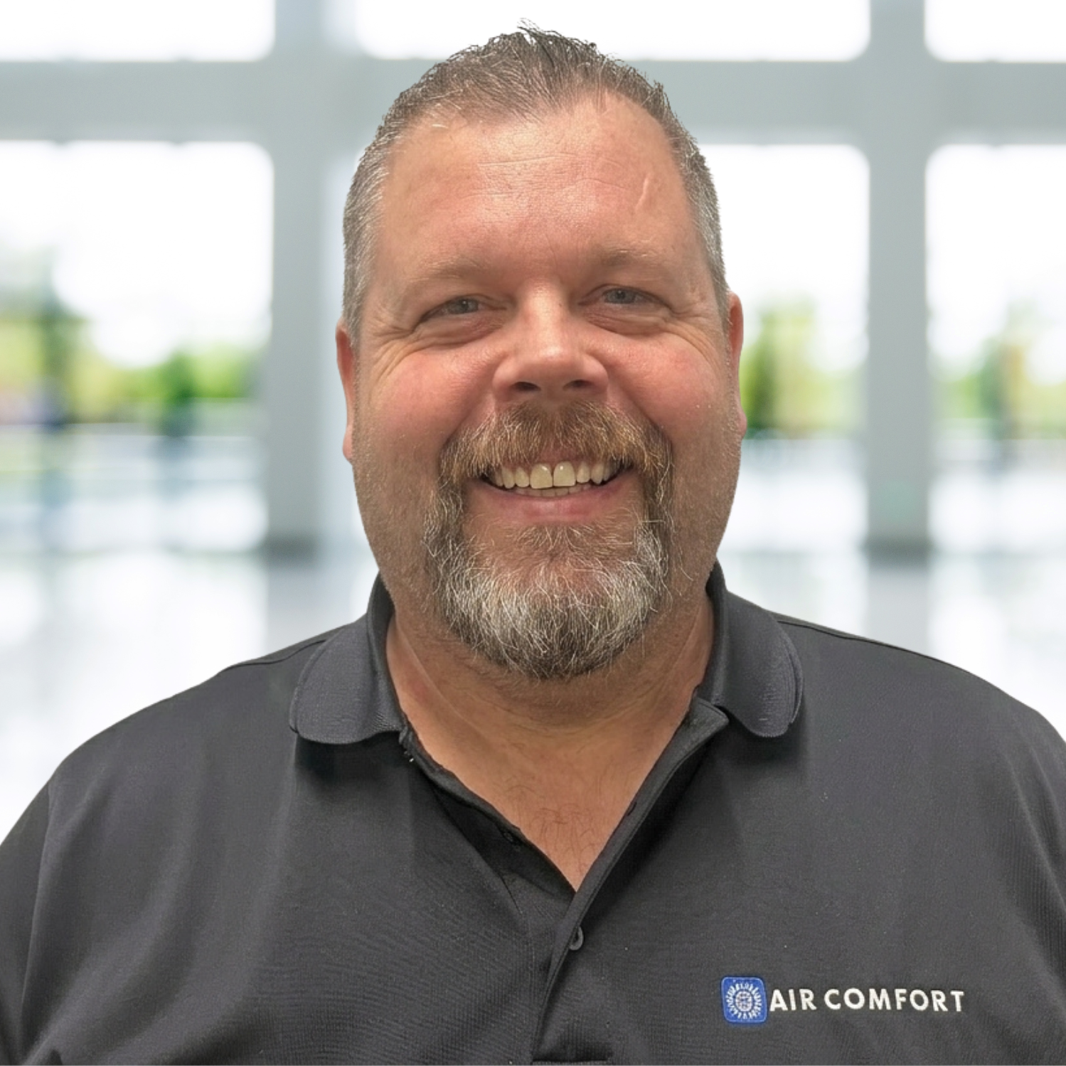 Smiling man in a dark polo shirt with an Air Comfort logo, standing in front of a bright blurred office background