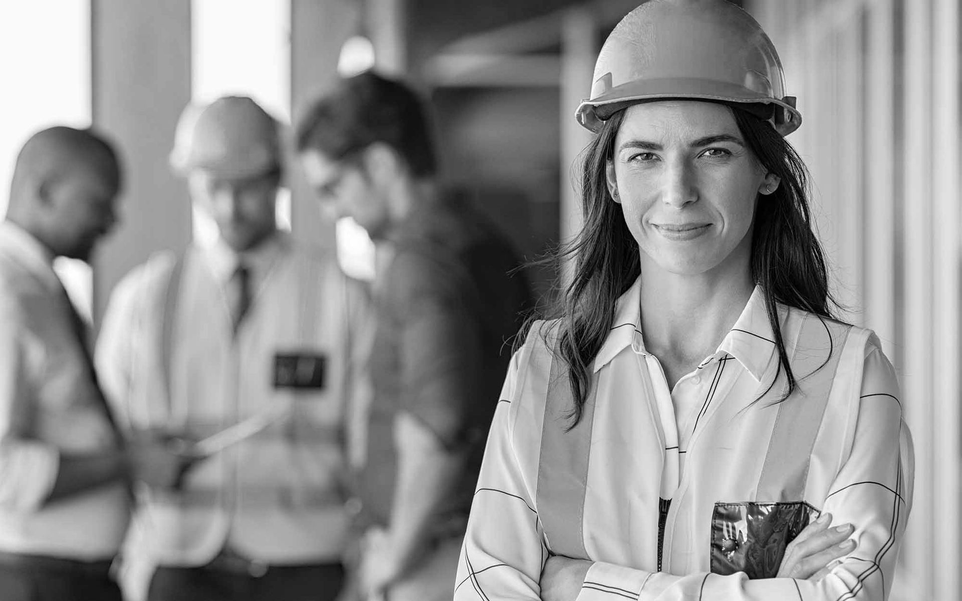 Engineer in a hard hat stands in an industrial workspace, looking at the camera with arms crossed.
