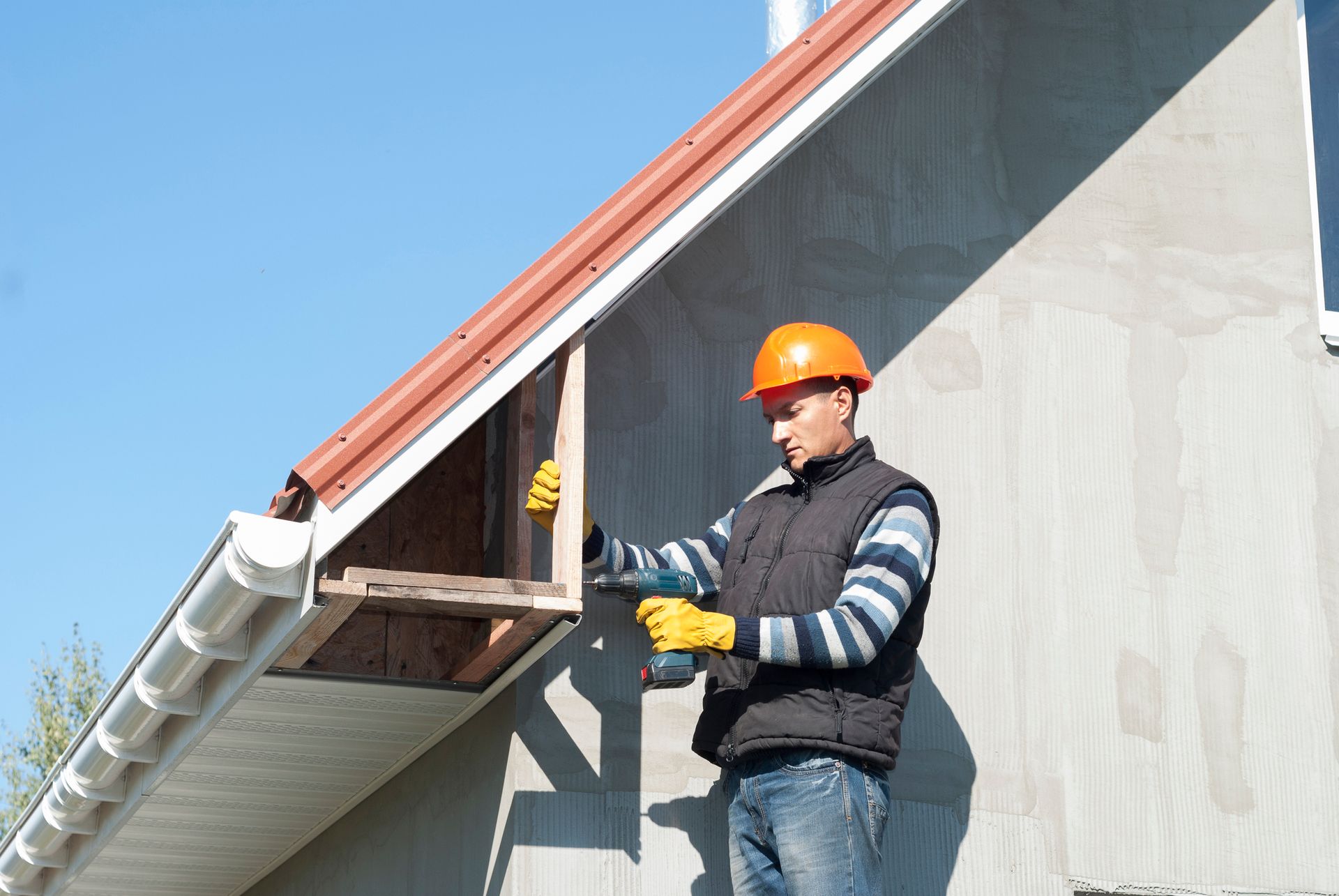 A male roofing contractor wears a safety hat and gloves as he drills a part of a roof’s fascia.