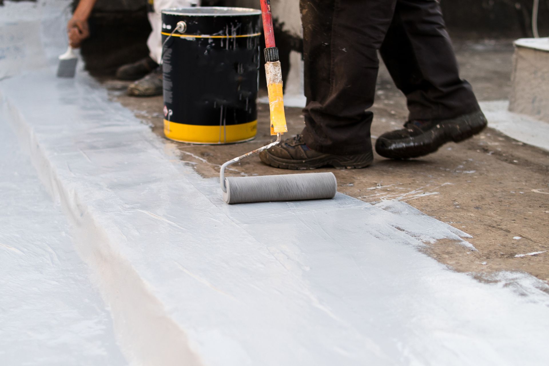 Person rolling white sealant on a roof, near a paint can, on a cloudy day.