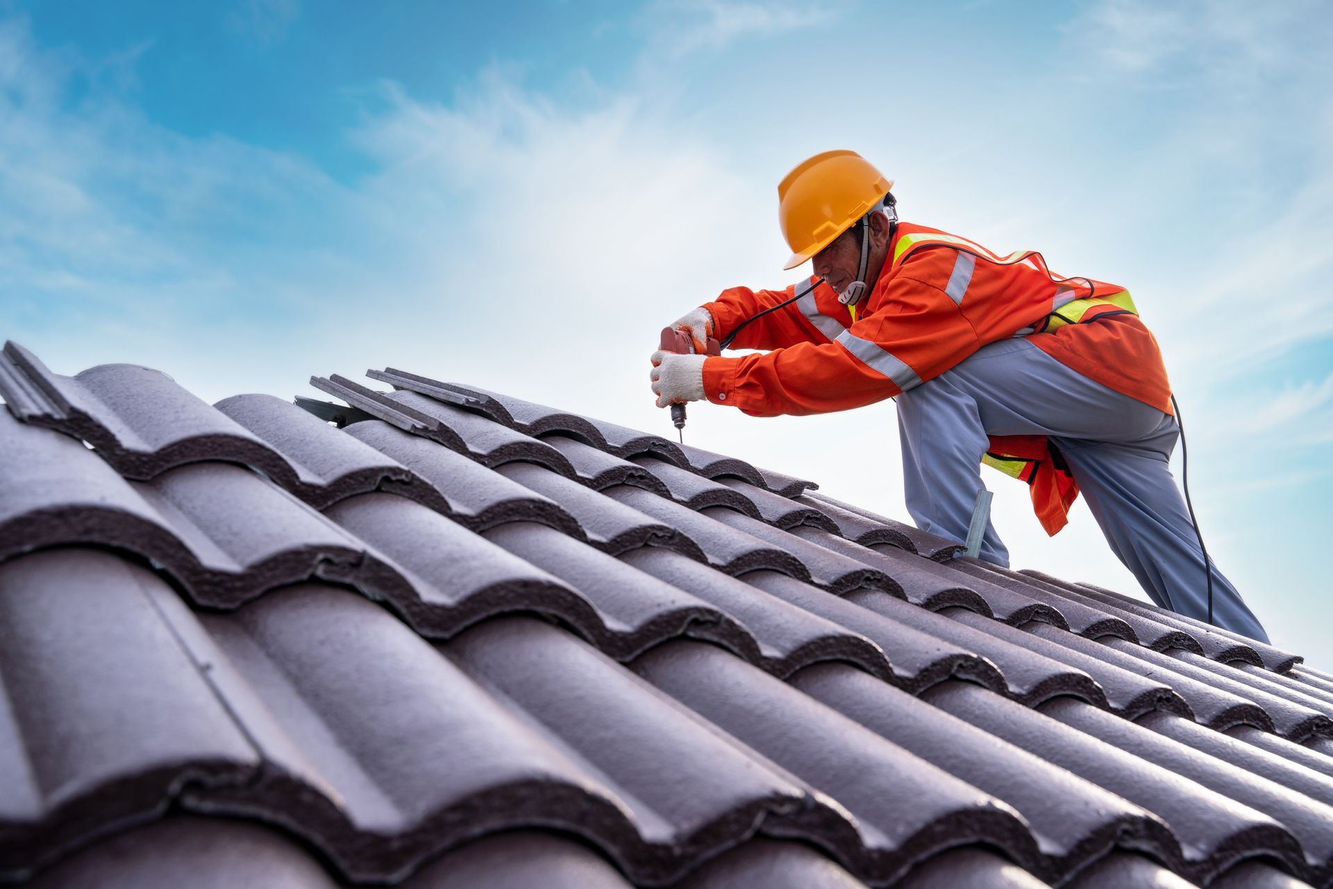 Roofer in orange safety gear installing dark brown tiles on a roof against a blue sky.