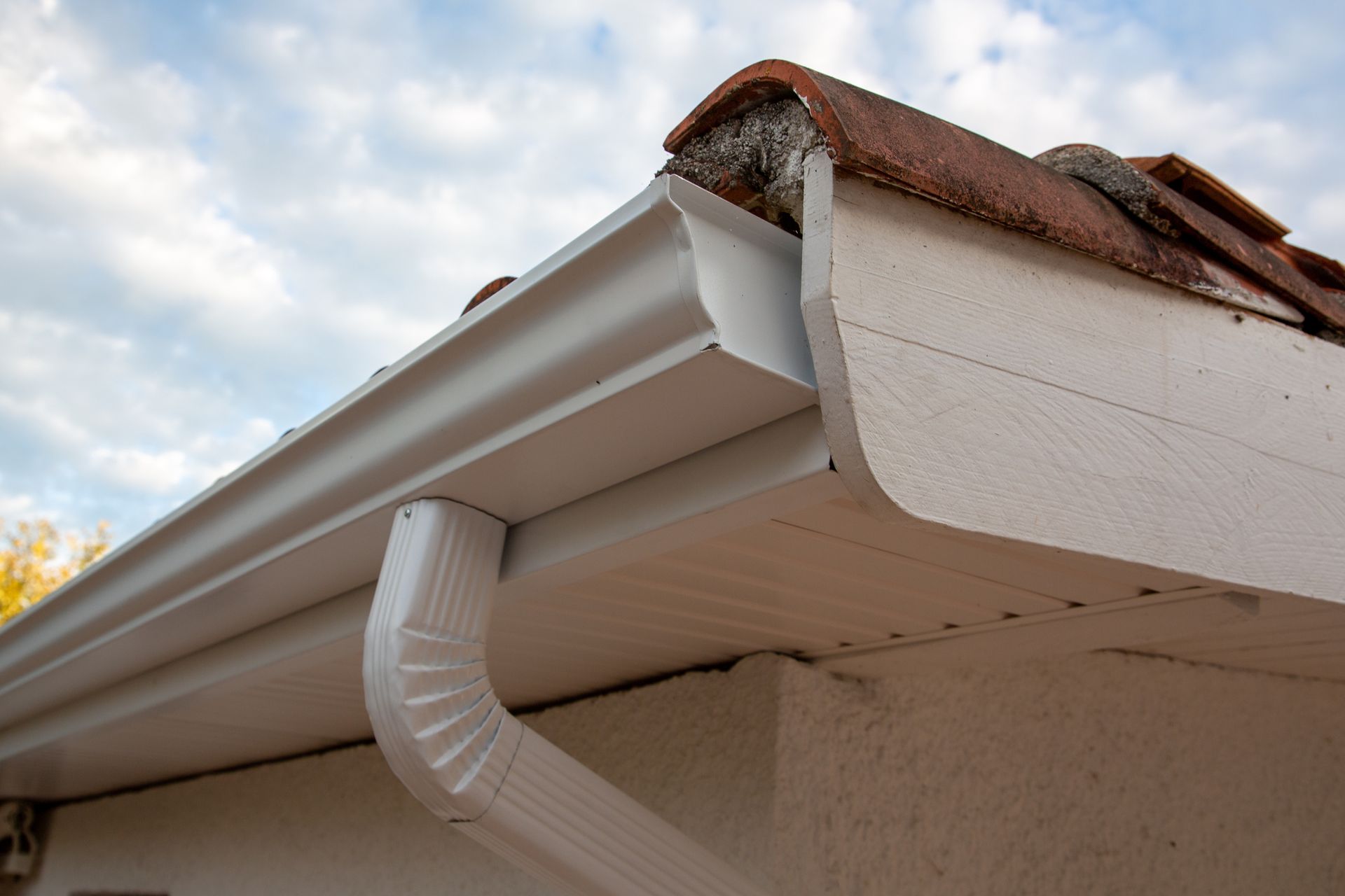 White rain gutter and downspout attached to a white-walled building with a brown-tiled roof.