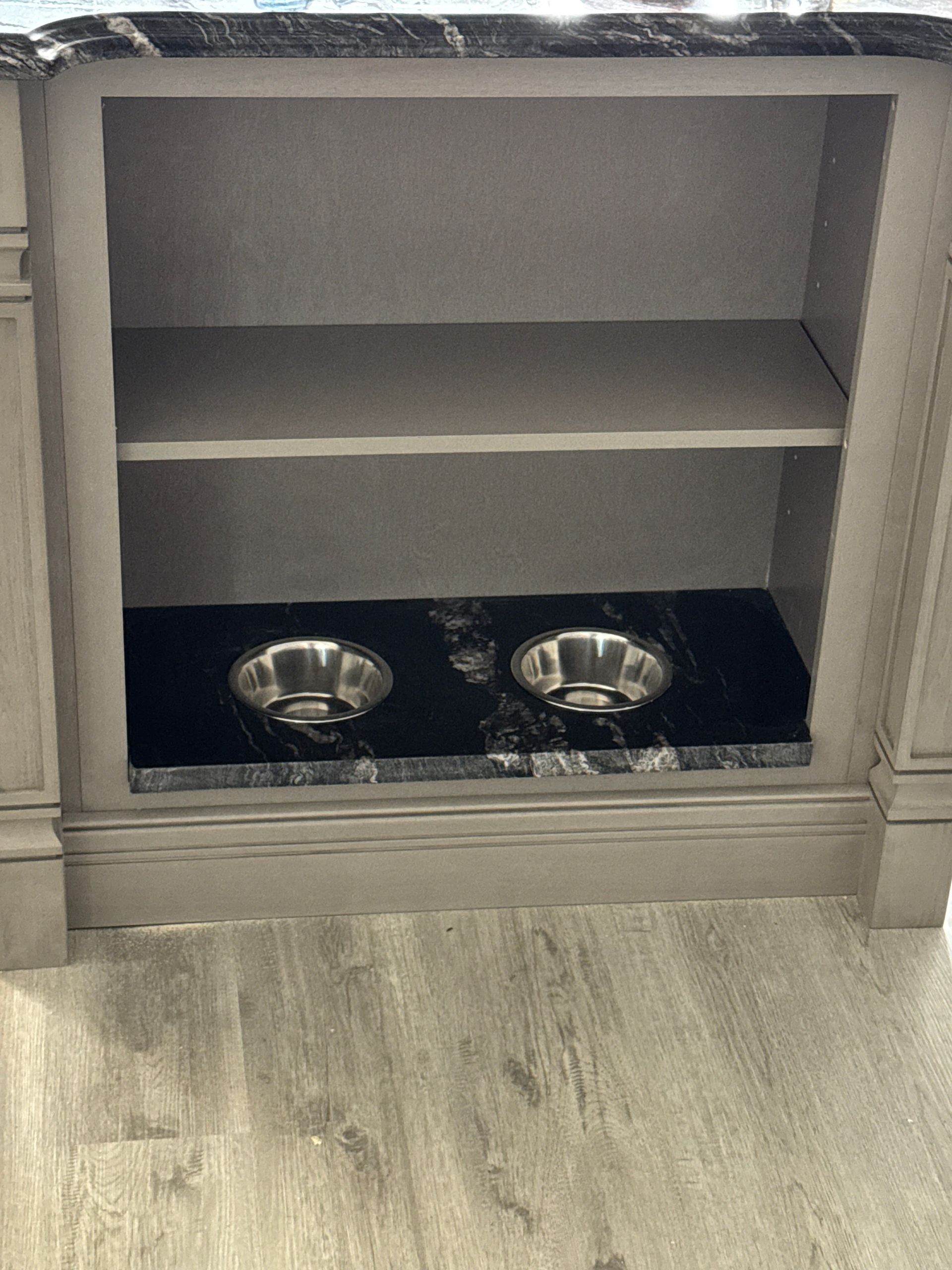 Built-in dog food bowls in a cabinet. Two silver bowls sit in a granite base. Gray cabinet with shelf above.