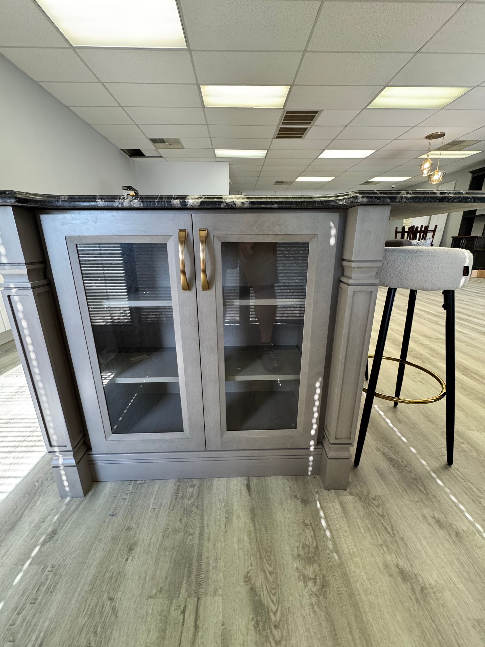 Gray cabinet with glass doors, gold handles, granite countertop, bar stool, and wood flooring in a store.