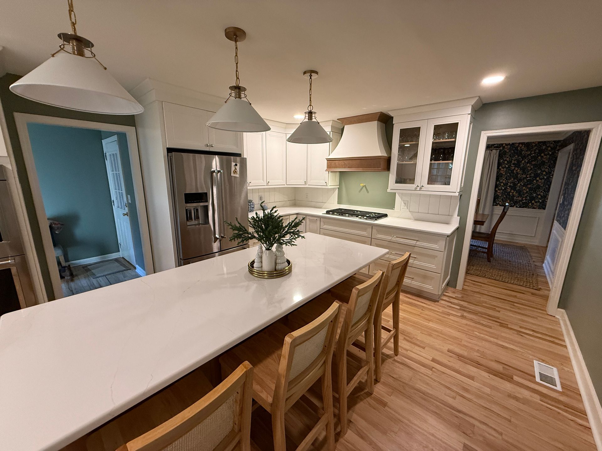 Kitchen with white island and countertops, light wooden cabinets and floors, and three pendant lights.