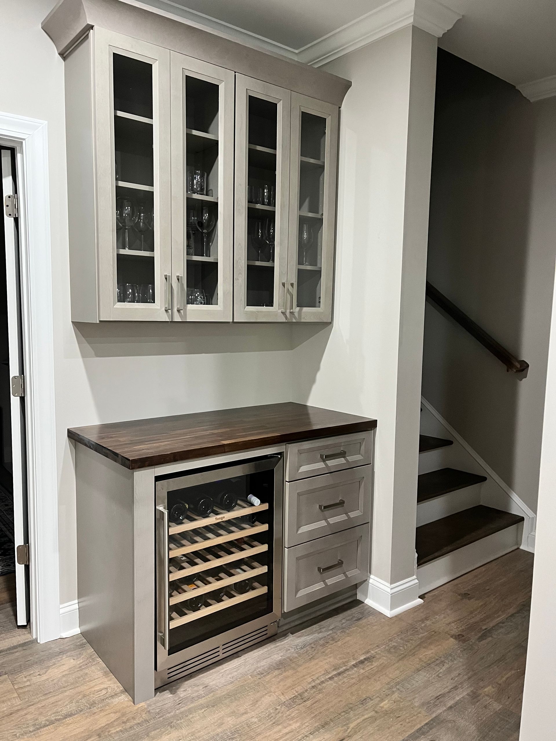Built-in gray bar cabinet with wine cooler, drawers, and upper glass-door cabinets; next to a staircase.