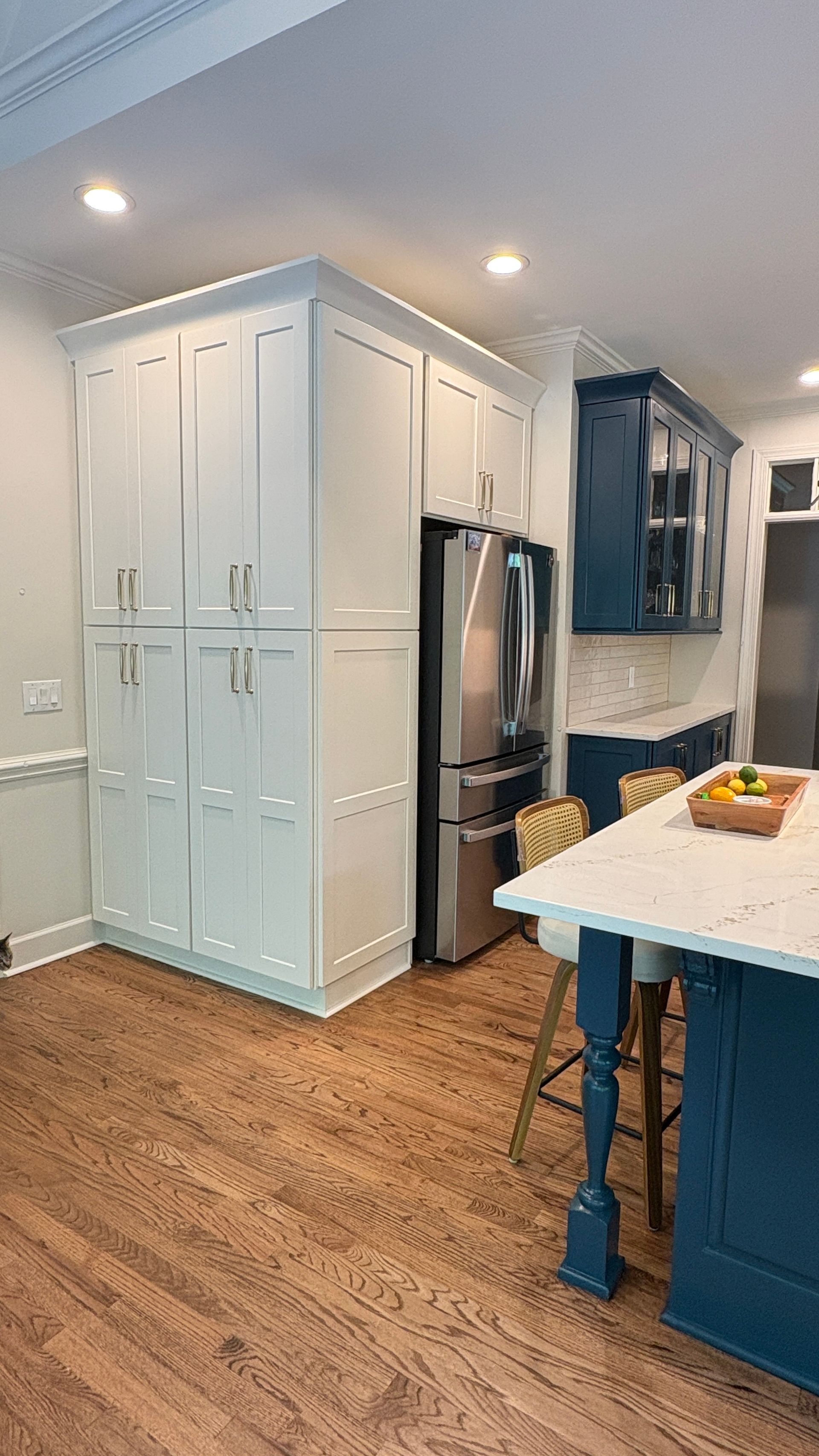 White and blue kitchen with wood floor, stainless steel refrigerator, and island with stools.
