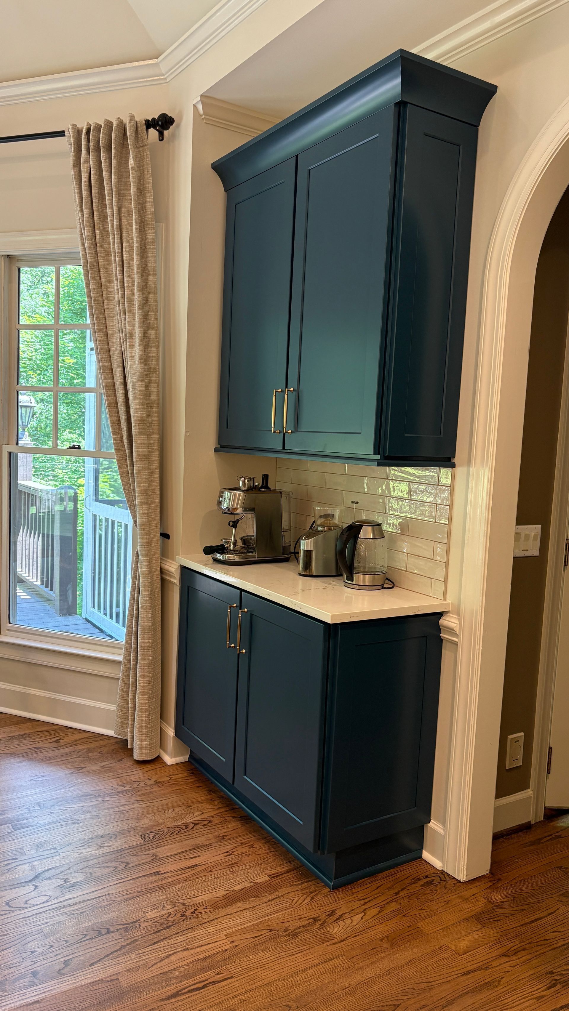 Built-in navy blue cabinets with appliances on a white countertop, next to a window with beige curtains.
