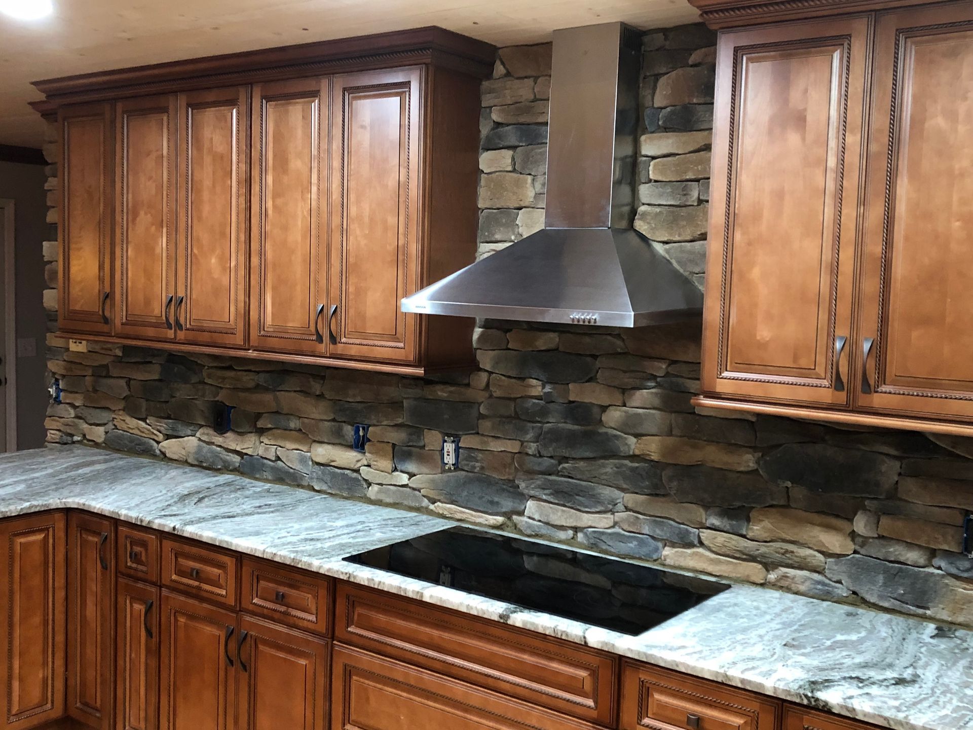 Kitchen with brown cabinets, stone backsplash, and granite countertop. Stainless steel range hood above cooktop.