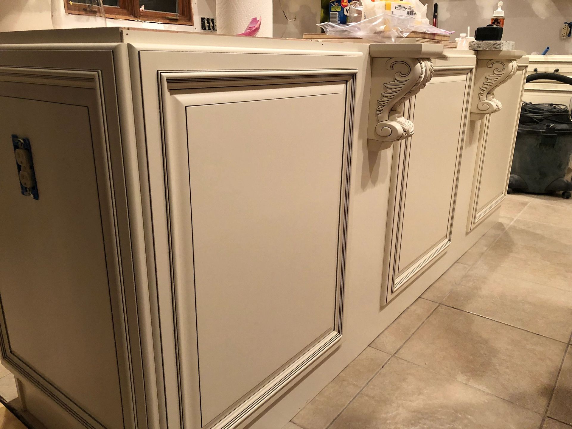 Cream-colored kitchen island with ornate trim and corbels, on a tiled floor, under construction.