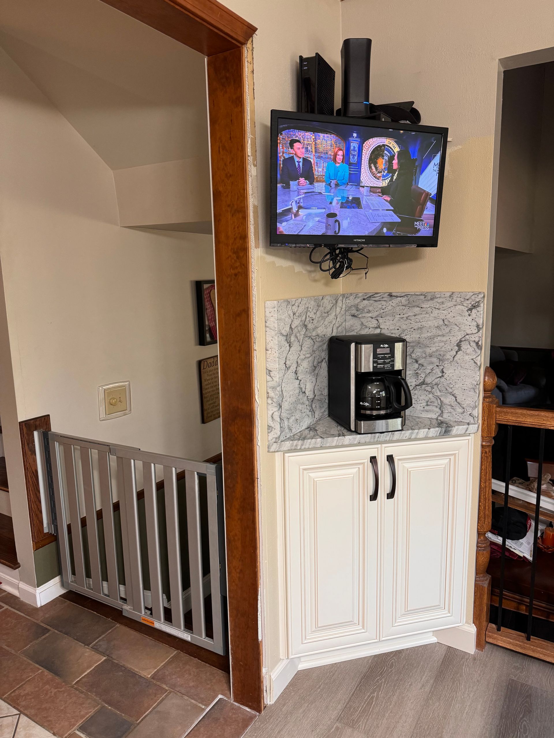 Coffee station with TV above. White cabinet, granite countertop, black coffee maker. Wooden trim and staircase.
