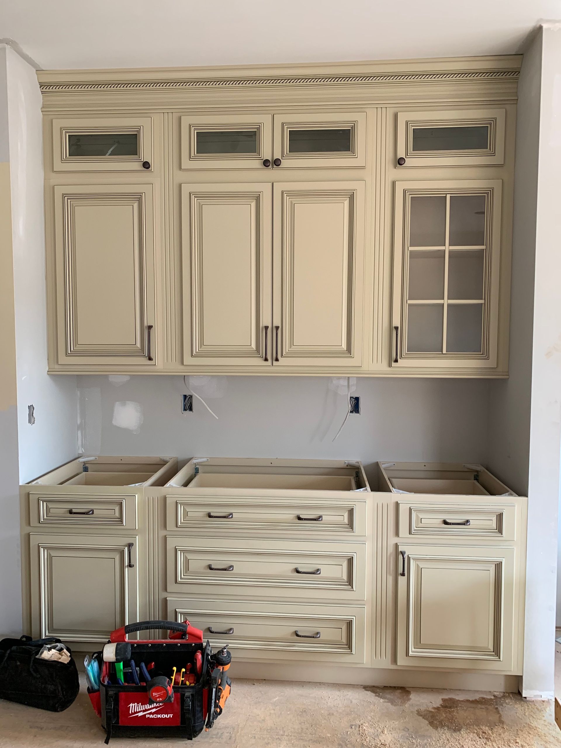 Cream-colored kitchen cabinets with distressed edges, installed on a wall. A toolbox sits on the floor in front.