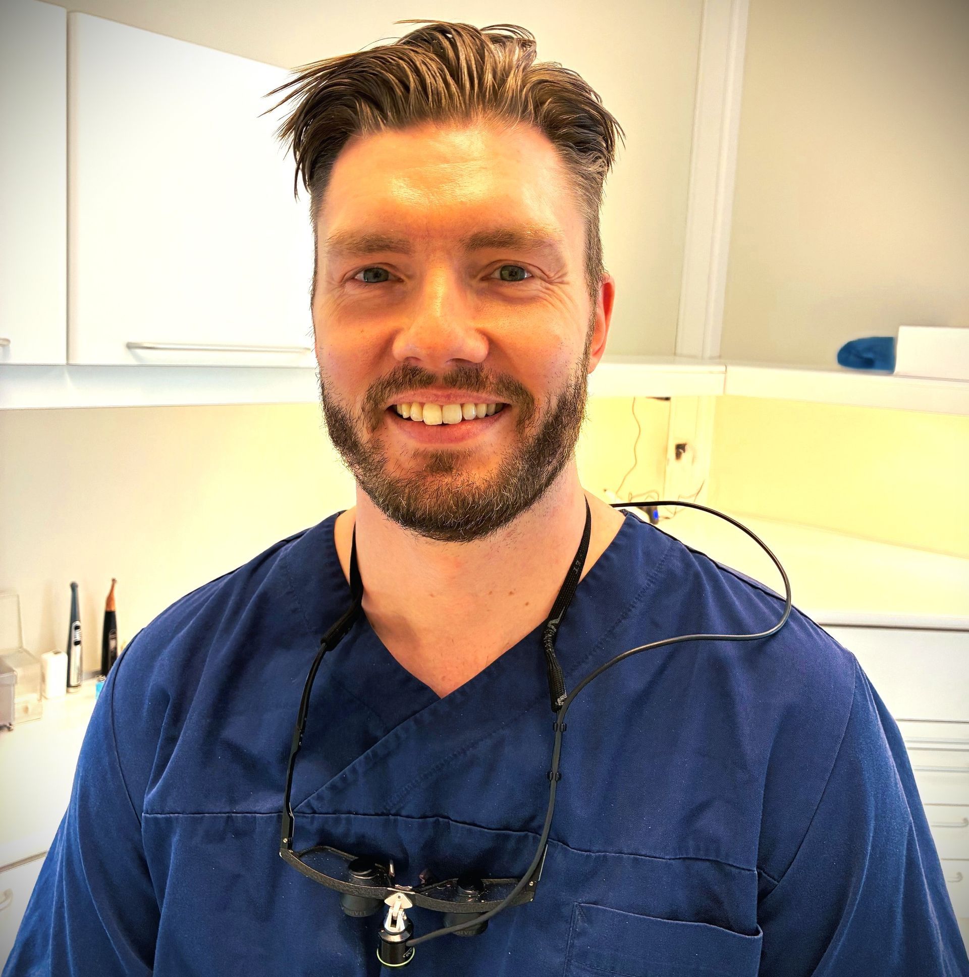 Tannlege Christian med briller og skjegg, iført blå skjorte og medisinsk maske, smiler til kameraet.