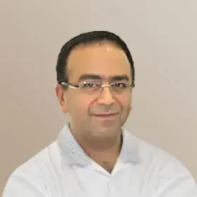 Tannlege Christian med briller og skjegg, iført blå skjorte og medisinsk maske, smiler til kameraet.