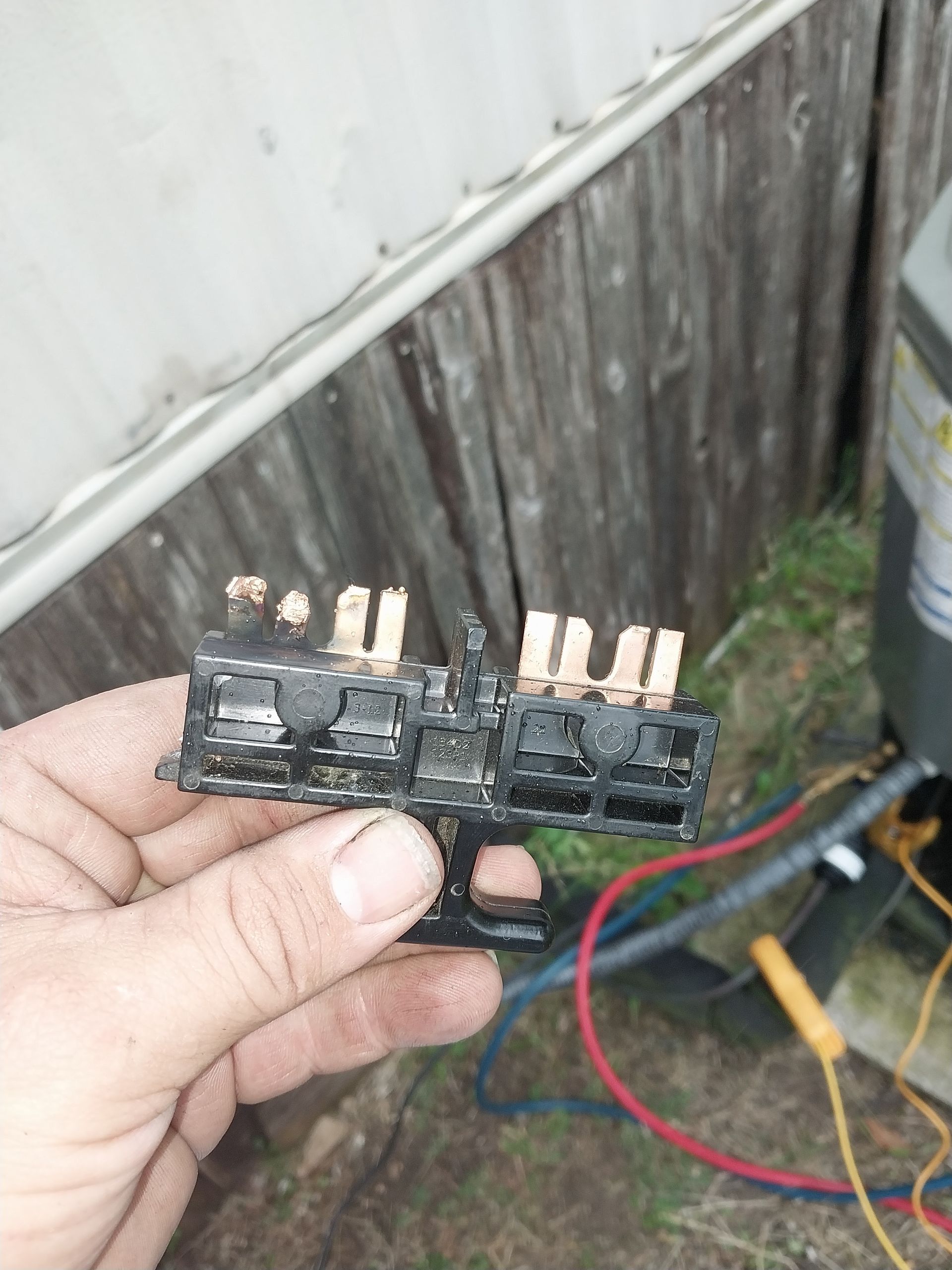 Hand holding a black electrical contactor with copper terminals, near wires and an air conditioner unit.