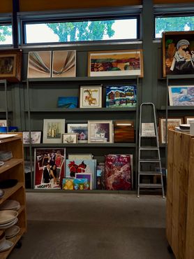 In een winkel staan ​​planken vol ingelijste kunstwerken; een ladder leunt tegen de muur.