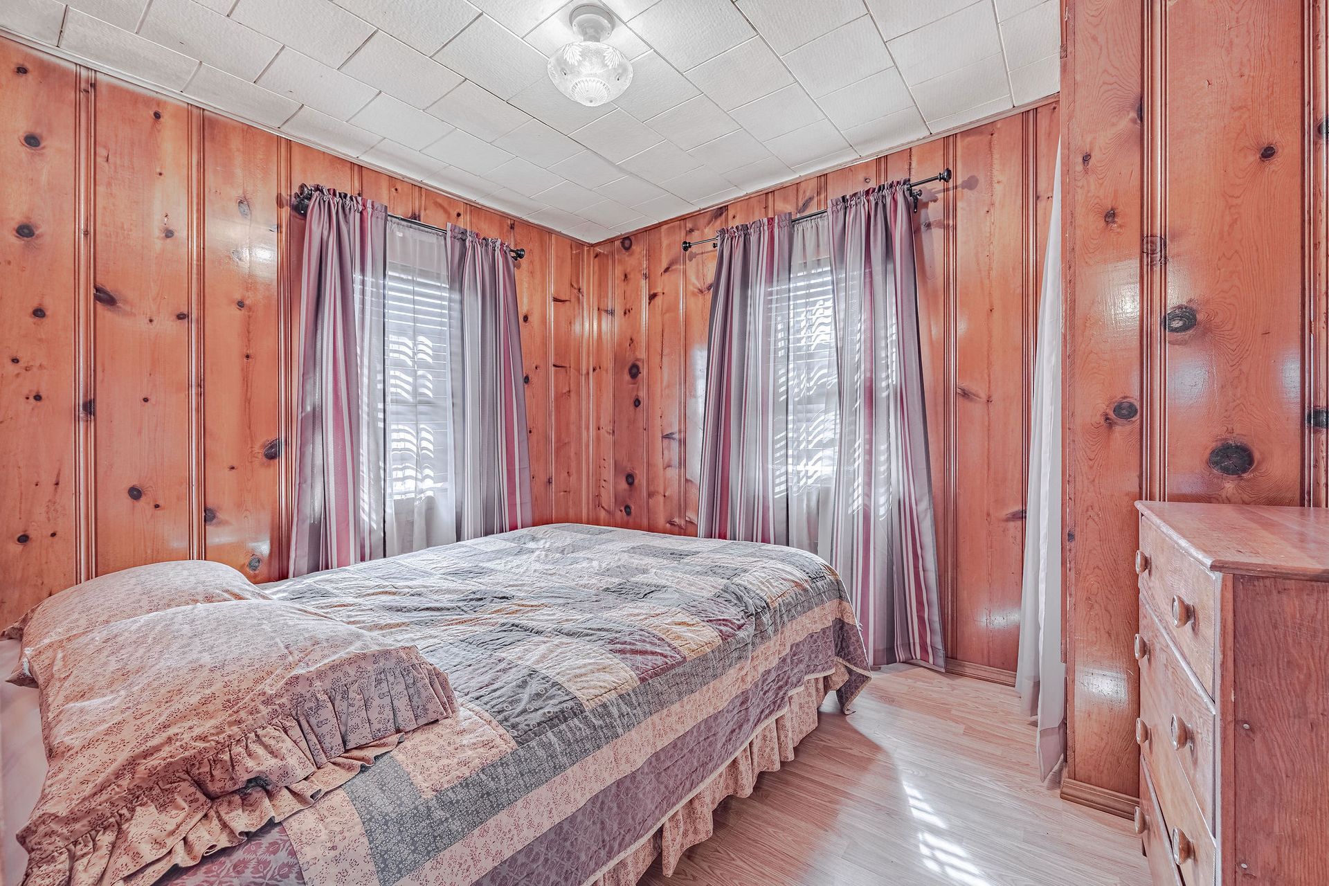 A bedroom with wood paneling , a bed , dresser and two windows.