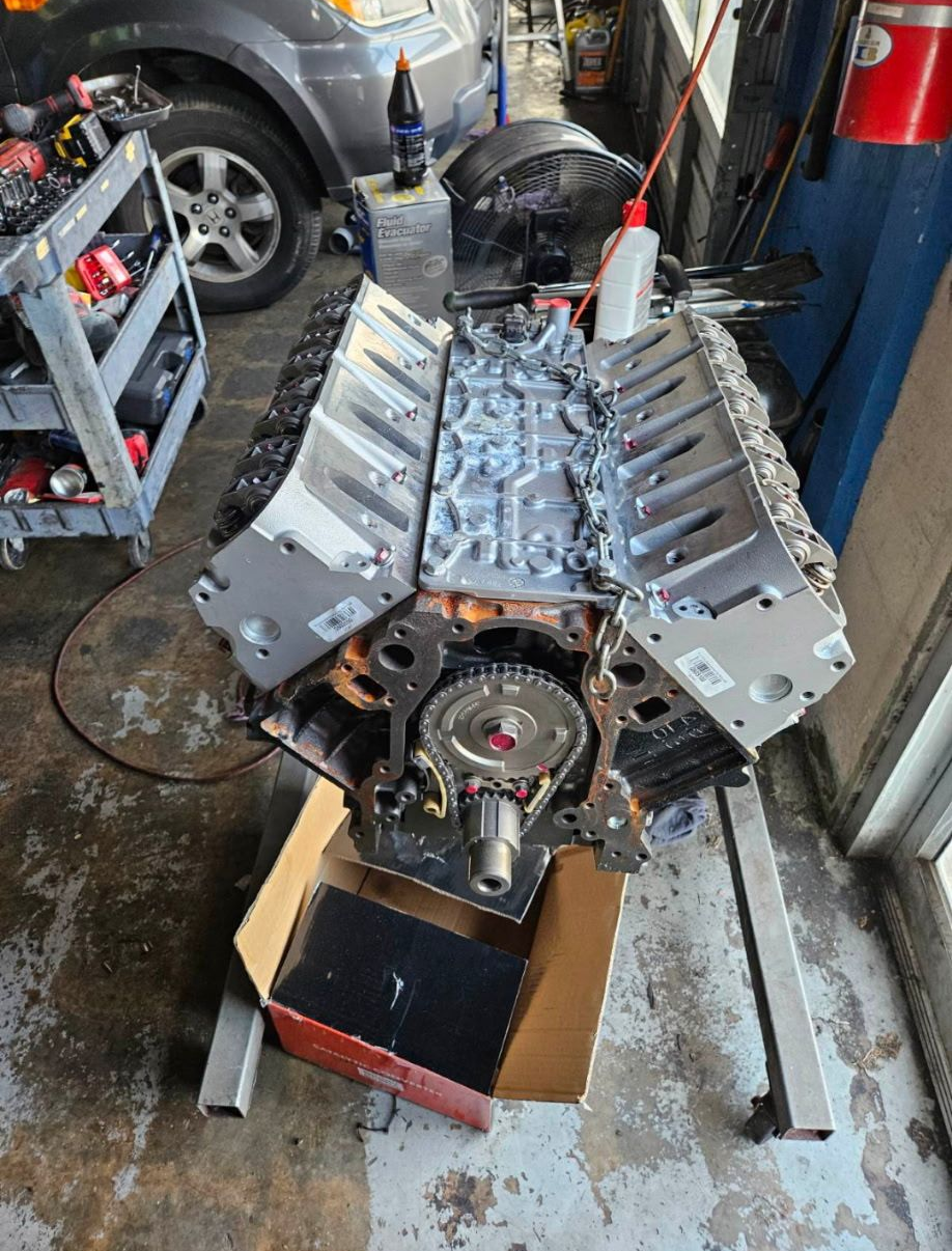 An engine block sitting on a stand, possibly under repair. Garage setting with car visible in background.