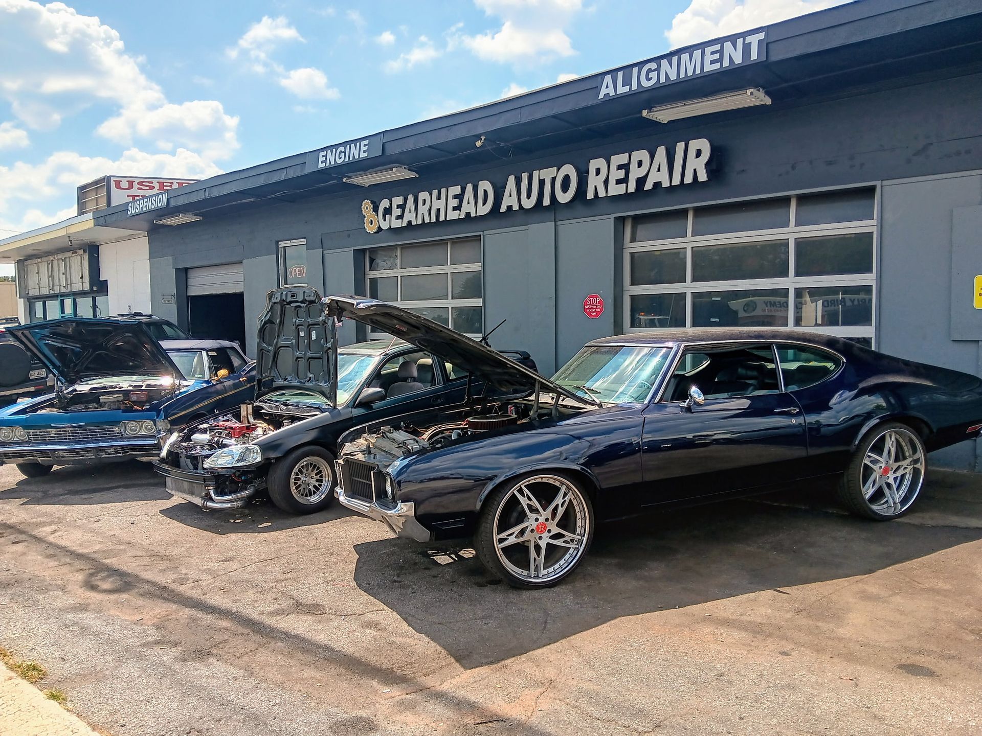 Classic cars parked outside a repair shop with open hoods. 