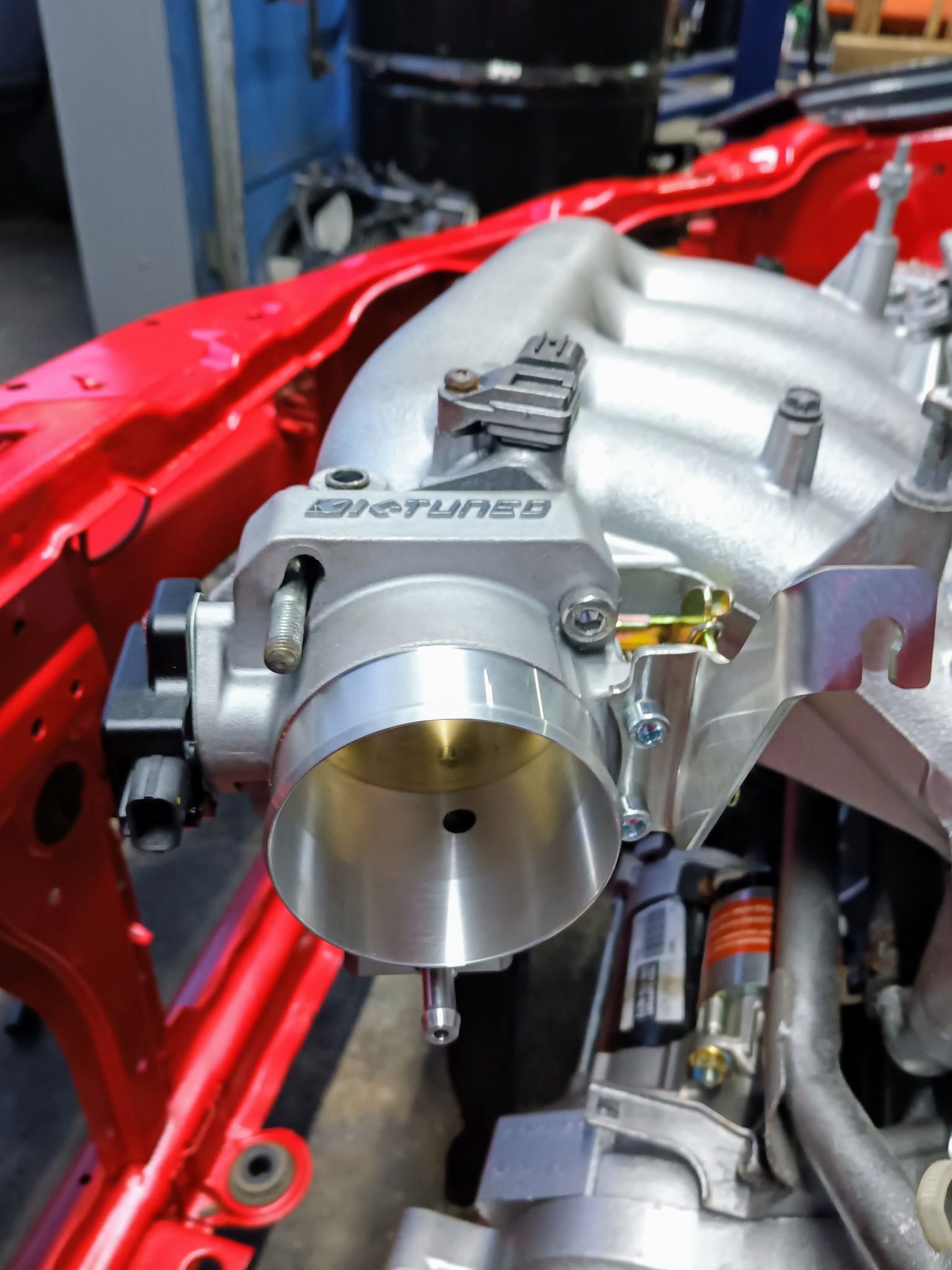 Close-up view of an engine's throttle body in a red engine bay. Silver throttle body is mounted on a manifold.
