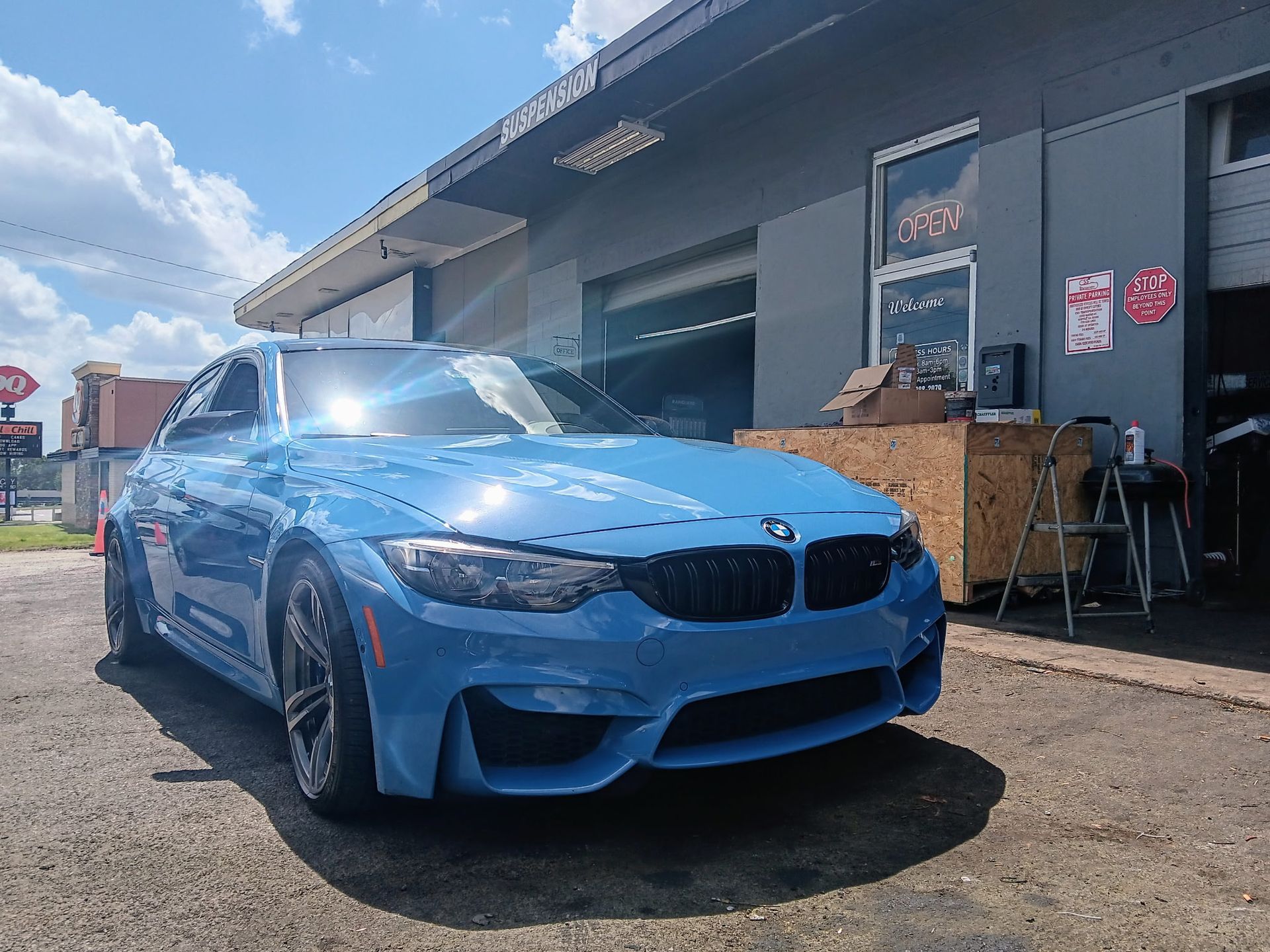 Blue BMW M3 sports car parked in front of a gray auto repair shop on a sunny day.