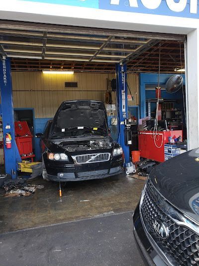 A car with its hood open inside a repair shop, car lift, tools, and a second car in the foreground.