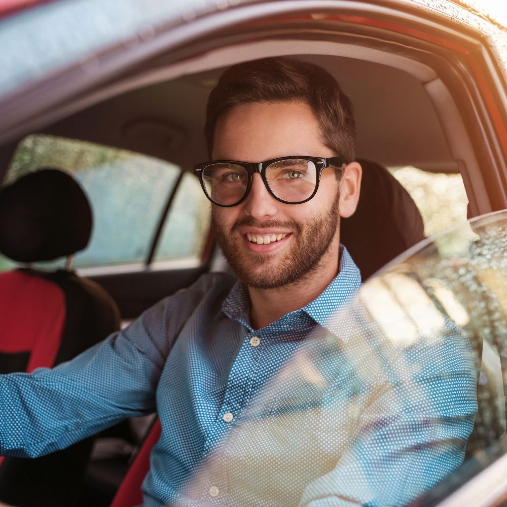Man wearing glasses smiles inside a car, looking directly at the viewer. Man in glasses smiling, sitting in a red car, looking at the camera.