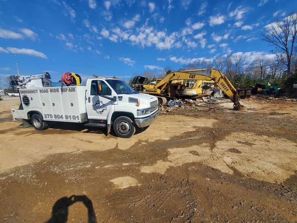 A white truck is parked in a dirt field next to a yellow excavator.