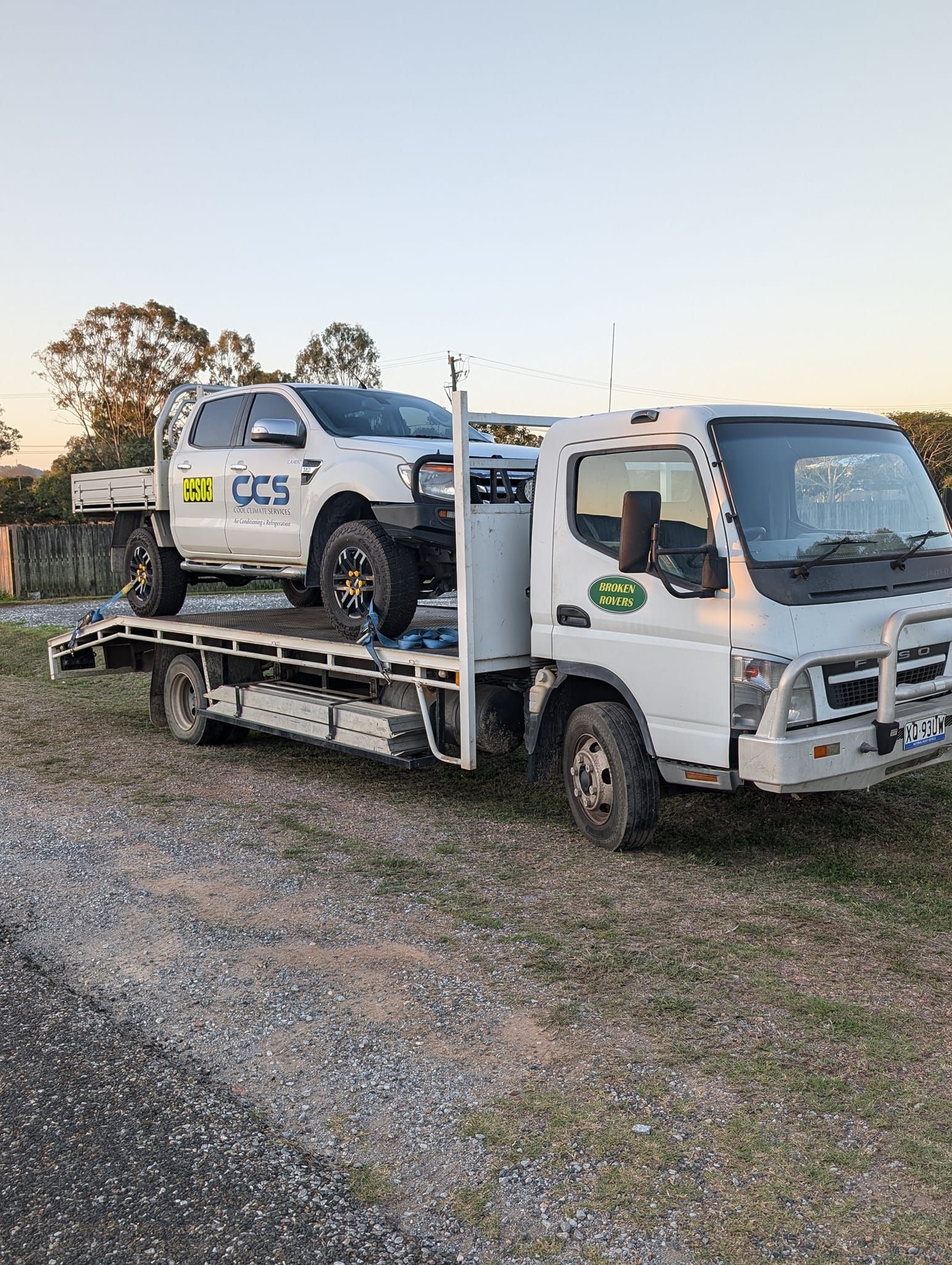 White tow truck carrying a white pickup truck on a gravel lot; daytime.