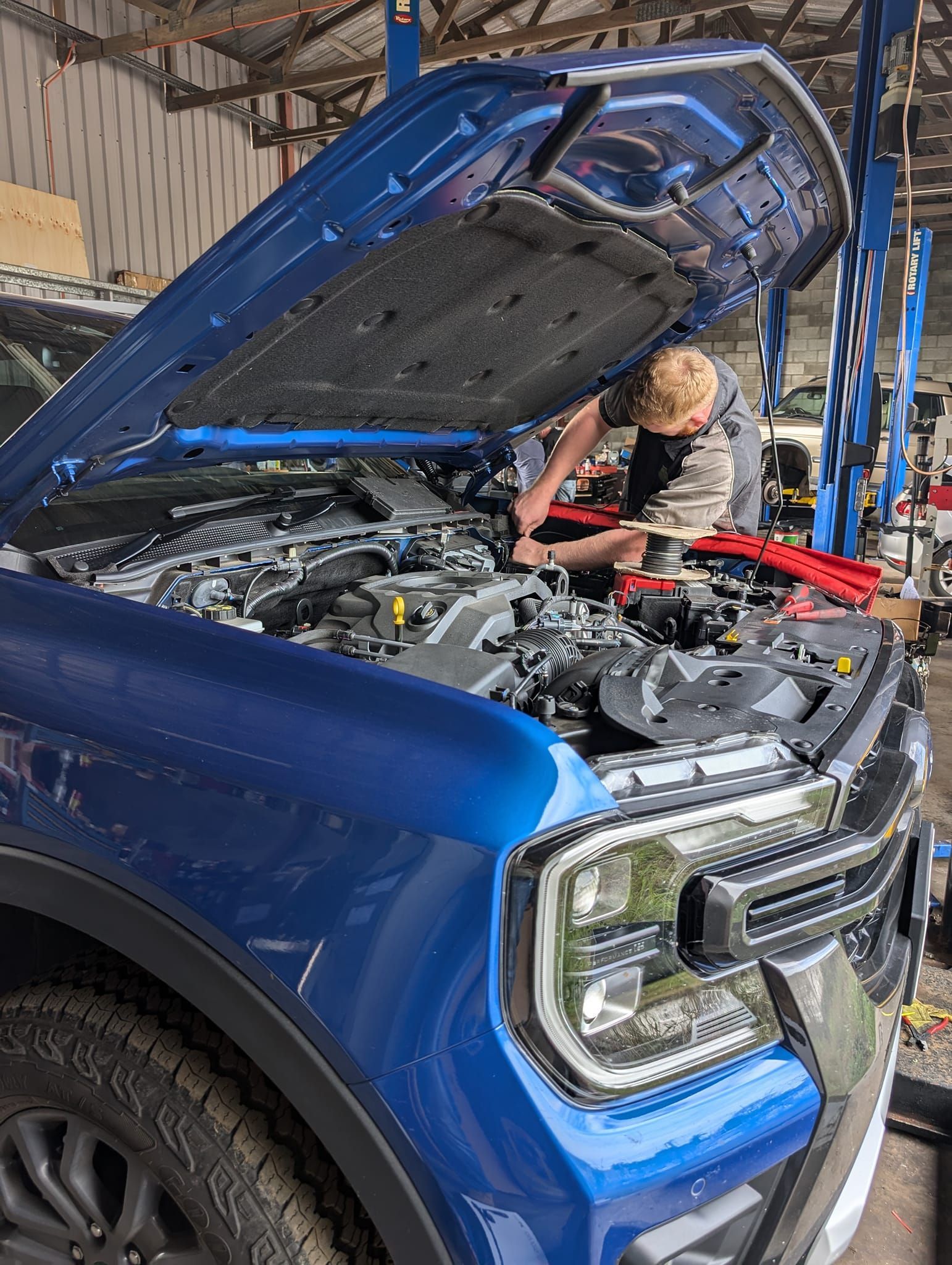 Blue SUV with hood open, mechanic working on engine in a garage.