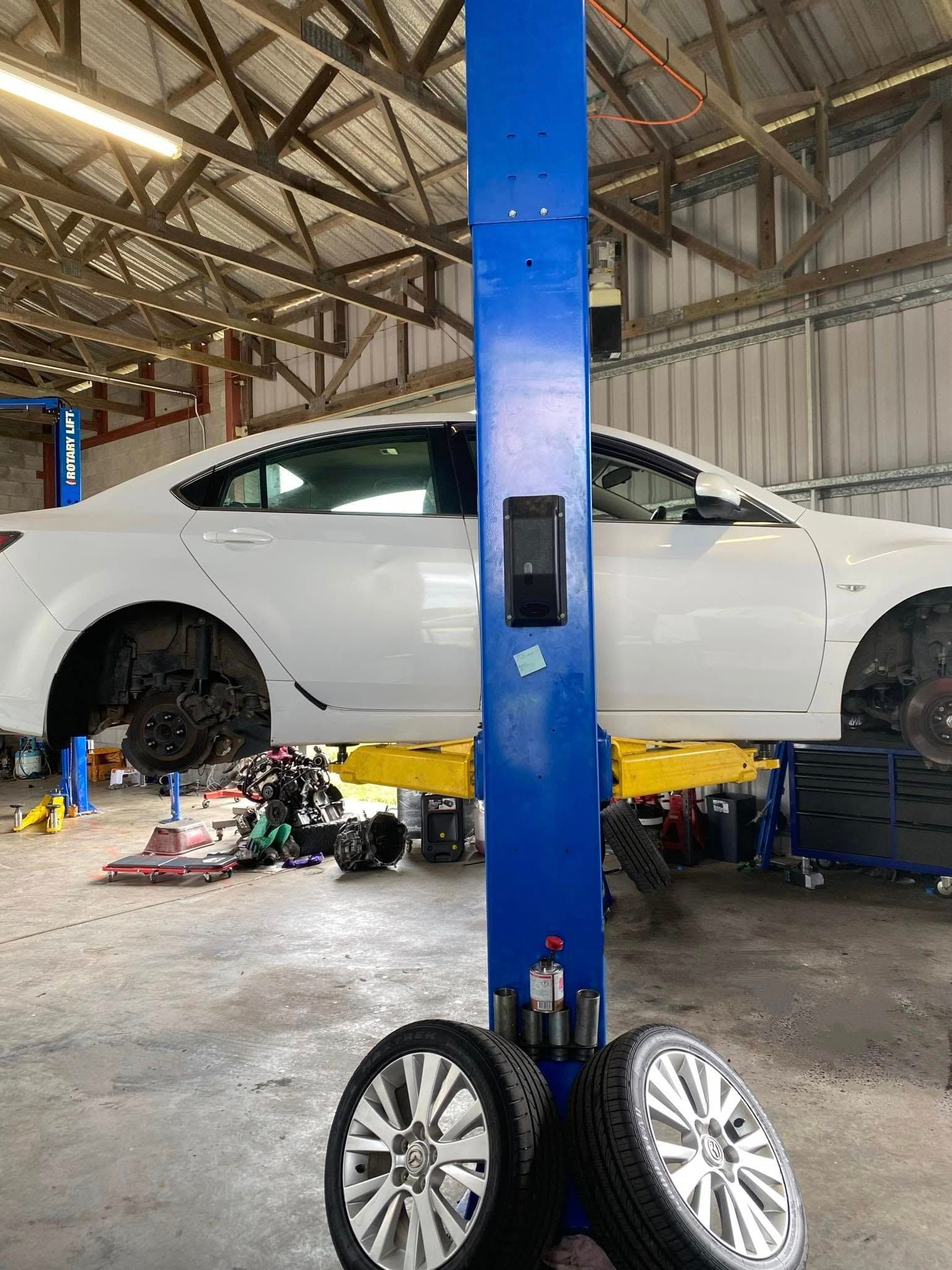 A white car raised on a lift in a garage. Two tires rest on the floor; blue lift column in foreground.