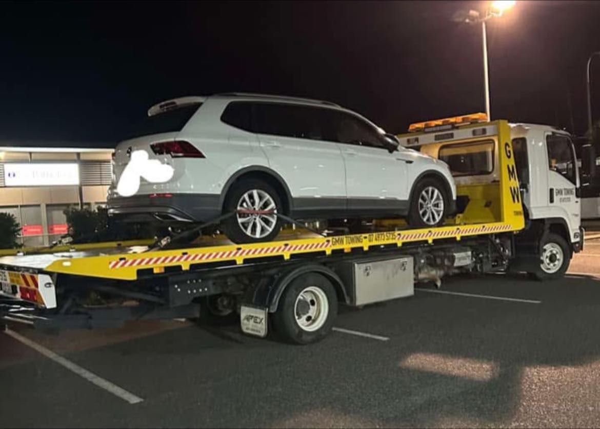 A small excavator is secured on a flatbed tow truck with yellow detailing, parked on green grass.