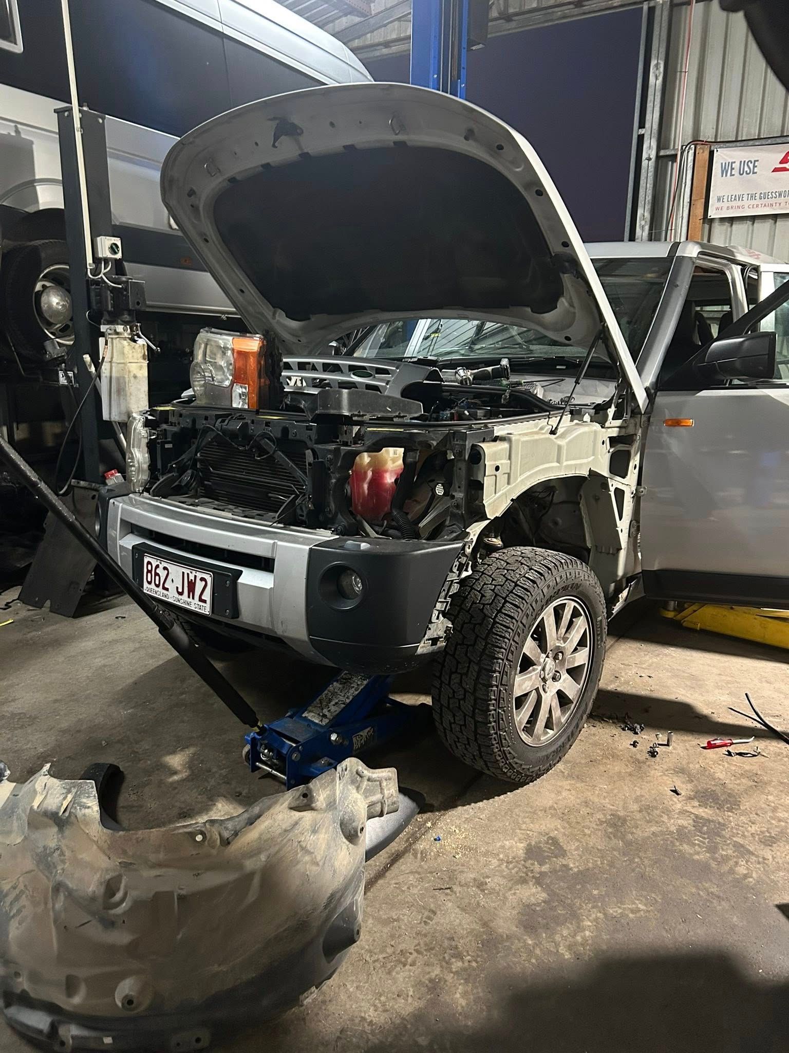 SUV in a repair shop with the hood open, front end disassembled, and a removed part on the floor.