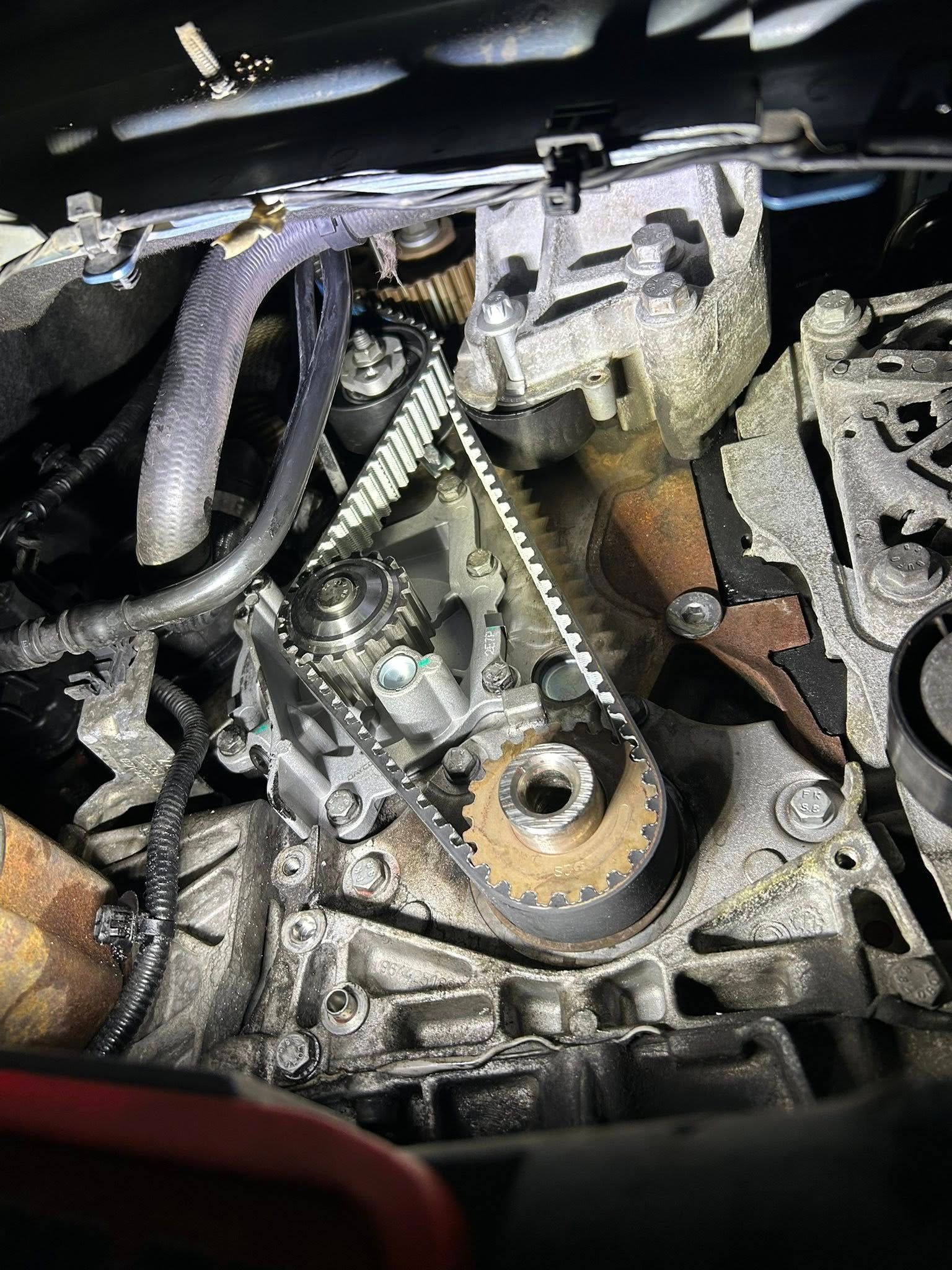 Engine components, including a timing belt, gears, and hoses, are visible under a car hood.