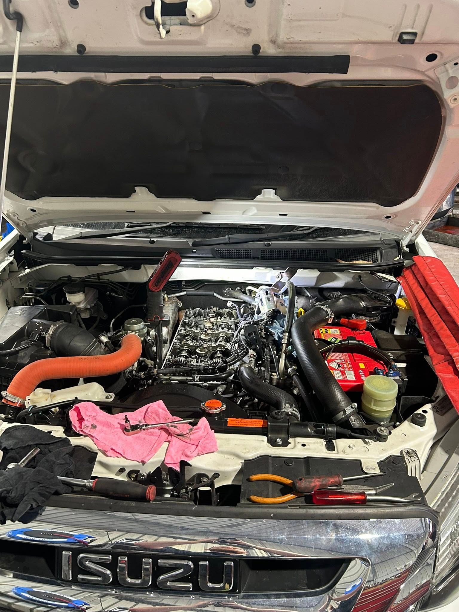 Yellow tow truck carrying a white caravan on a grassy area, trees in the background, sunny day.