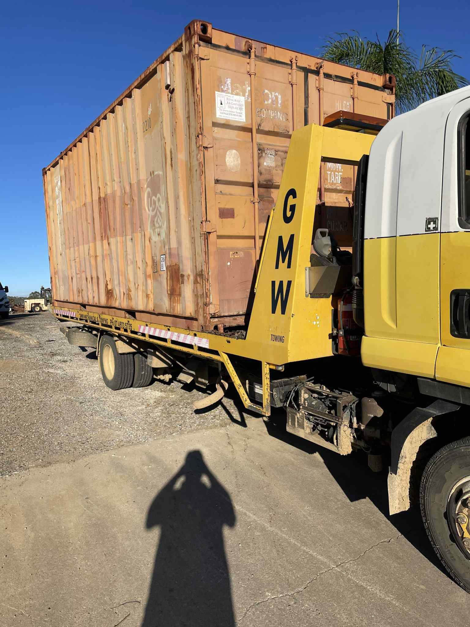 Yellow truck transporting a rusty brown shipping container on a paved lot. Shadow of photographer visible.
