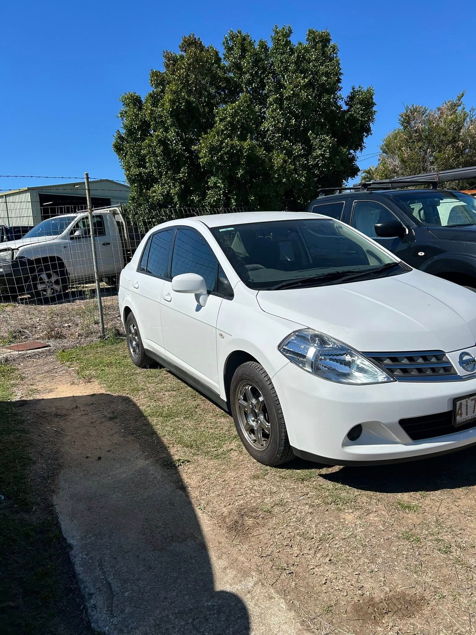 White Nissan Tiida parked outdoors on a sunny day. A green tree and other vehicles are in the background.