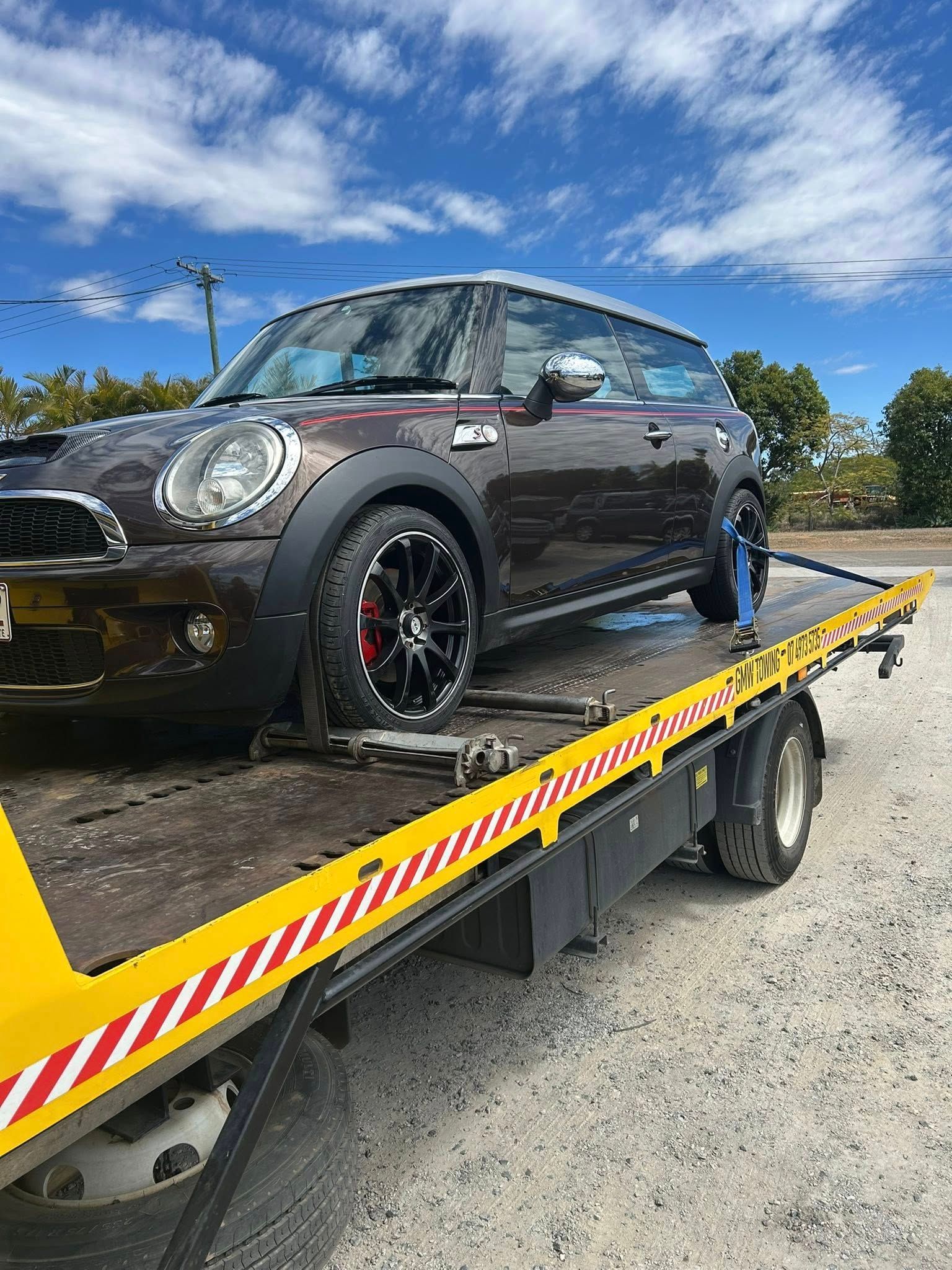 Brown Mini Cooper car being towed on a flatbed truck on a sunny day.