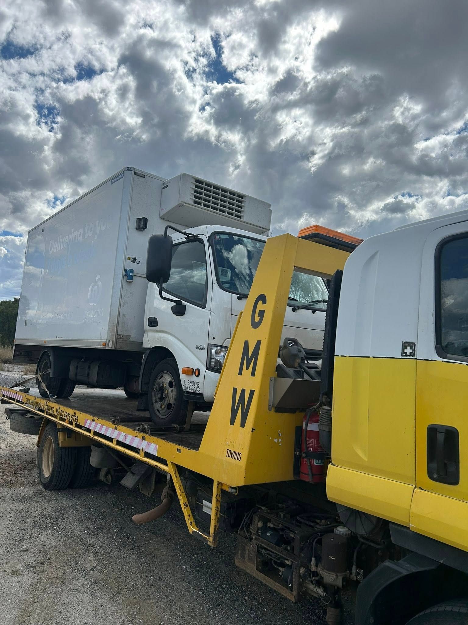 White refrigerated truck on a yellow tow truck bed under a cloudy sky. The tow truck has 