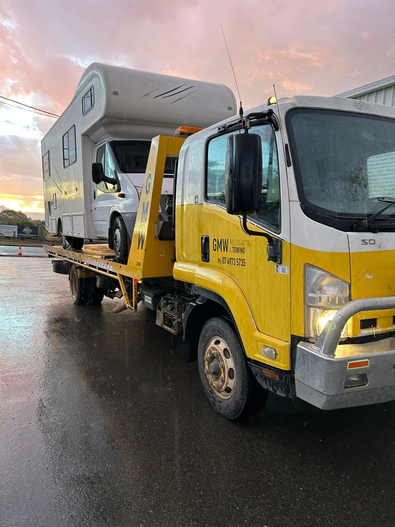 Yellow tow truck transporting a white camper van on a wet road at sunset.