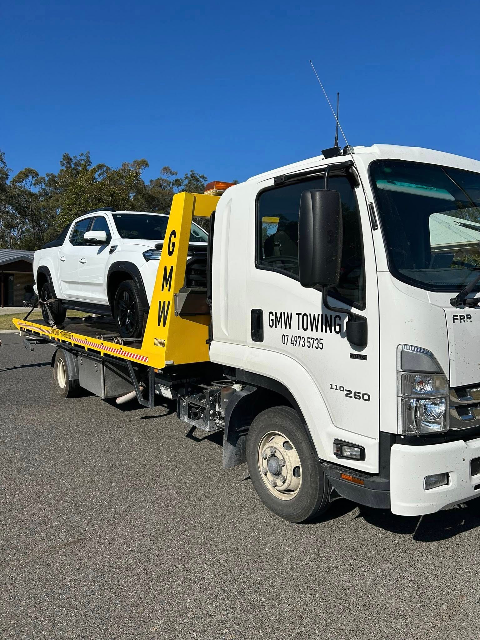 White tow truck carrying a white pickup truck on a gravel driveway under a blue sky.