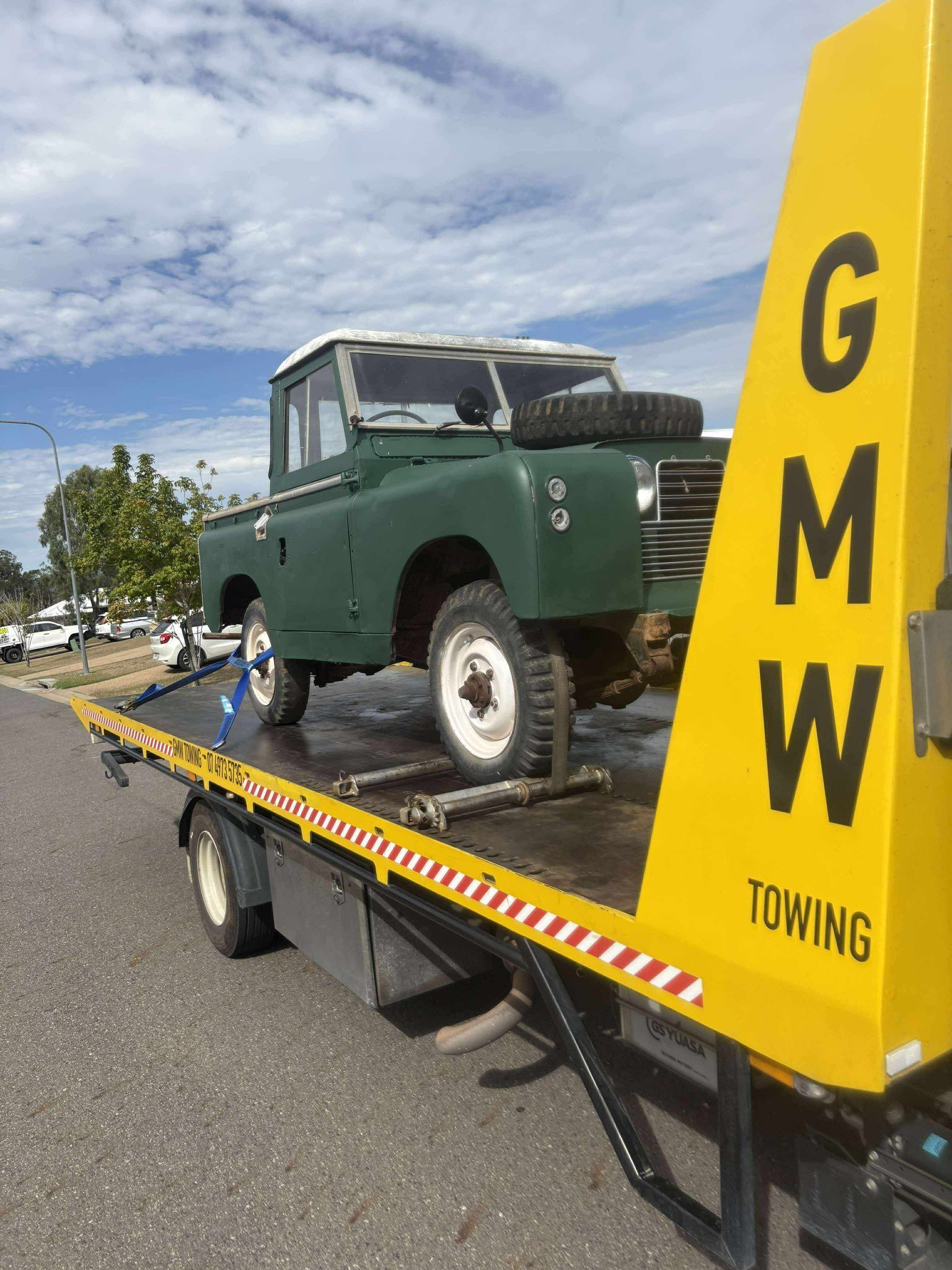 Green Land Rover on a flatbed tow truck. Yellow GMW towing sign. Cloudy sky.