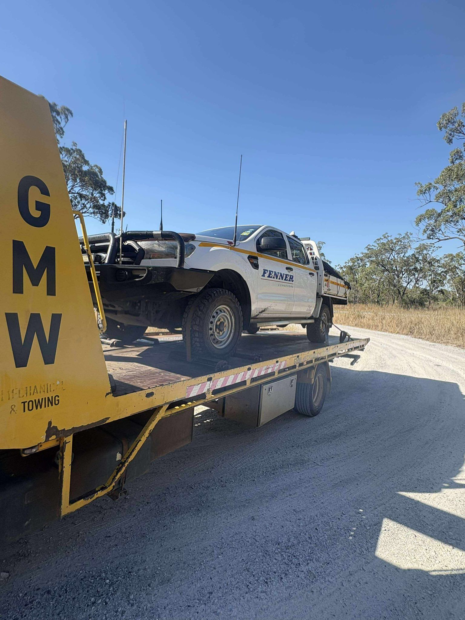 White pickup truck on a yellow flatbed tow truck on a gravel road, under a blue sky.
