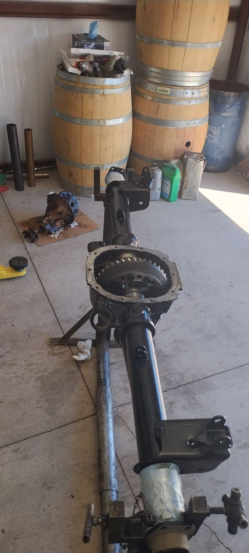 A car axle and differential assembly mounted on a stand in a workshop with wooden barrels in the background.